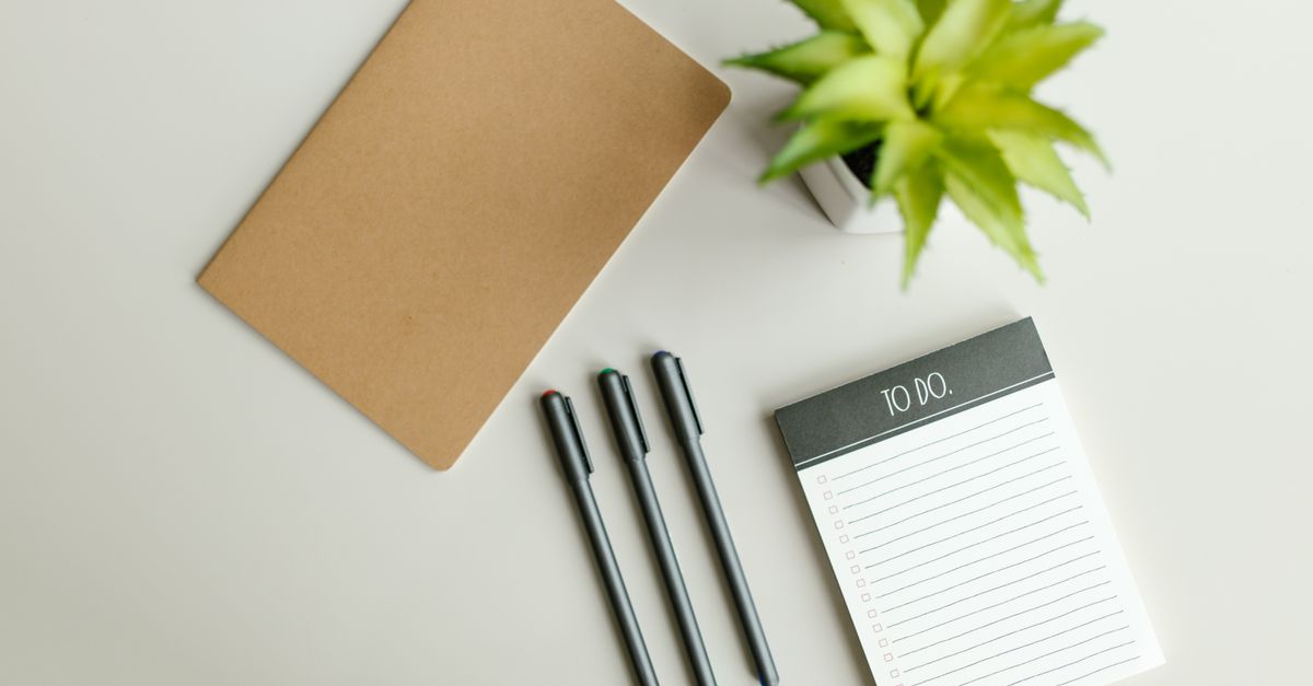 Flat lay of a to-do list, notebook, pens, and succulent on a light gray desk surface.